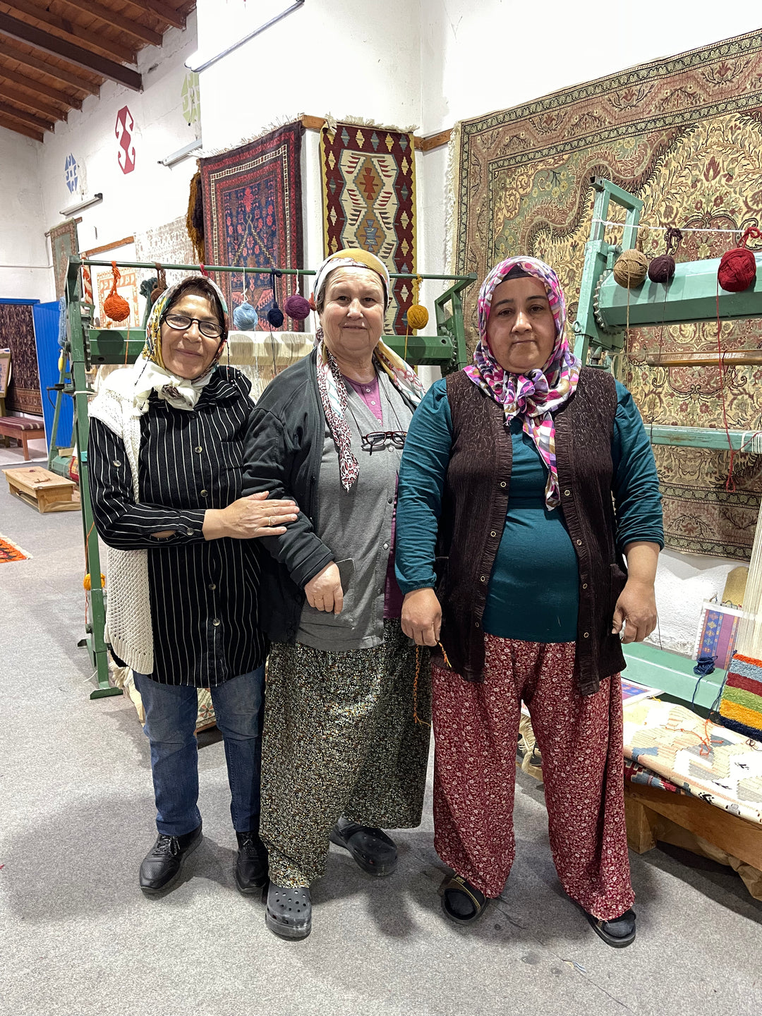 Three MaterialWorld.design artisans standing in a traditional rug workshop, surrounded by handwoven carpets and spools of yarn on looms