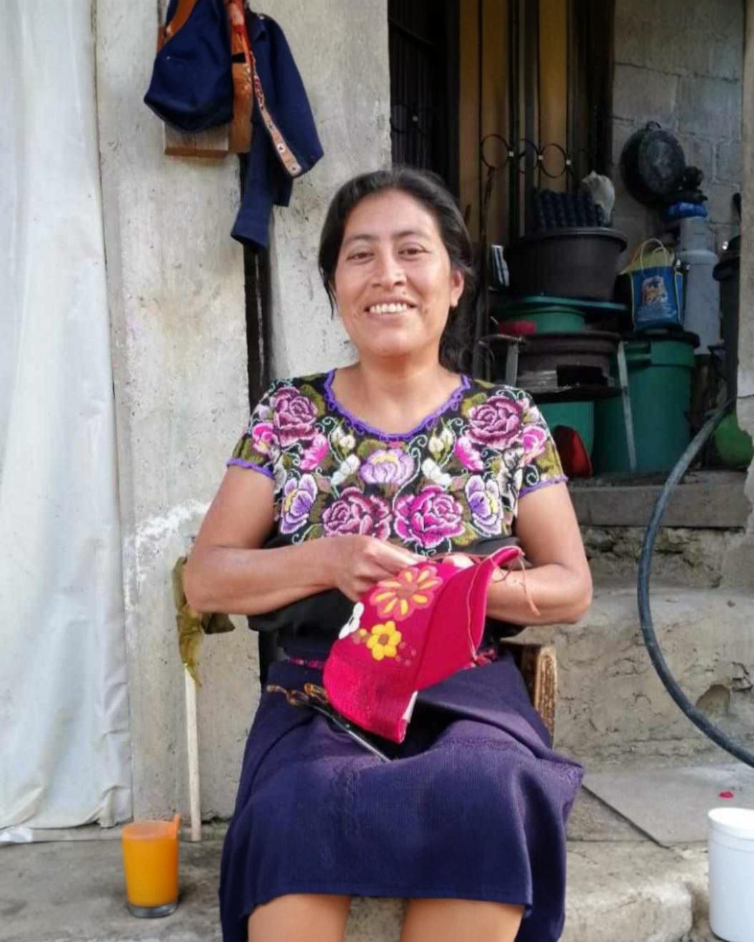 Smiling woman artisan seated outside, holding a pink fabric panel embroidered with yellow and red flowers
